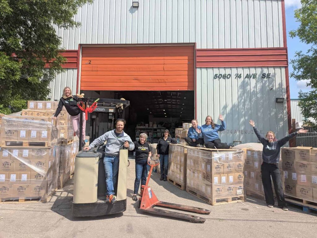 Pallets of Ziwi pet food donations with people celebrating the donation.